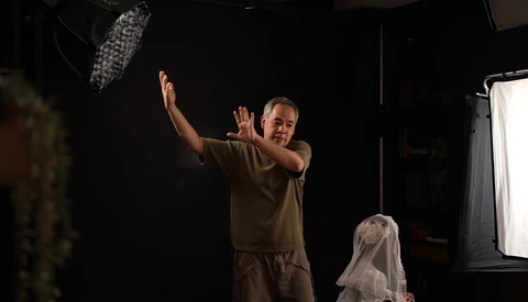 Director gesturing with hands raised during film production on a dark set with professional lighting equipment visible.