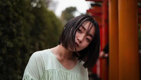 Young woman with short dark hair wearing a light green top, tilting her head with a contemplative expression against a blurred background with red and orange architectural elements.