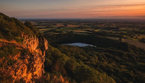 Dramatic cliff face illuminated by golden sunset light, overlooking forested valley with distant lake and agricultural plains.