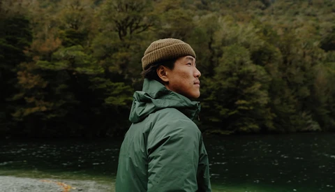 Person in green jacket and tan beanie looking toward a forested shoreline beside still water.
