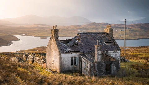Dilapidated stone cottage on moorland with mountains and lochs in background at golden hour.