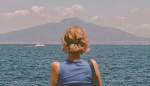 Woman with blonde hair viewed from behind, gazing at a distant mountain across blue water.