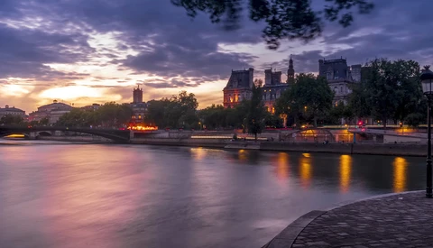 Illuminated riverside buildings at twilight reflected in still water under a dramatic purple and gold sky.