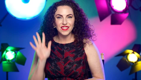 Woman with curly dark hair waving at camera against colorful studio lighting setup.