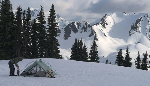Backpacker standing beside a dome tent on snow-covered slope with snow-capped peaks and evergreen forest in background.