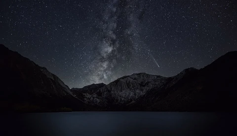 Milky Way galaxy stretches across a starlit night sky above snow-capped mountains and a dark alpine lake.