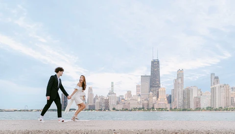 Couple in formal attire playfully running on a beach with a city skyline in the background.