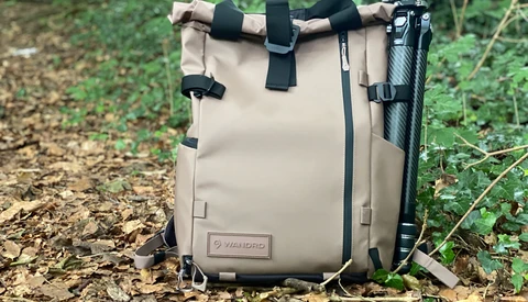 Tan canvas camera backpack with black straps and tripod attachment, photographed outdoors on forest ground.