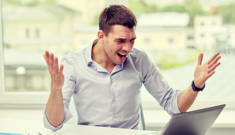 Man at desk with laptop gesturing expressively with both hands raised.