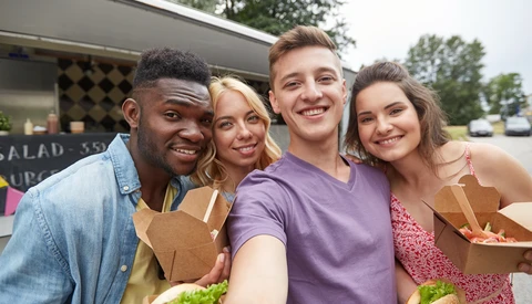 Four young adults smiling at a food truck window while holding takeout containers.