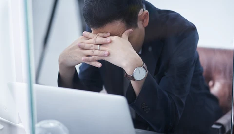 Man in dark blazer covering his face with both hands while seated at laptop, appearing stressed or fatigued.