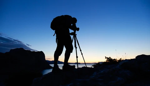 Silhouetted photographer with tripod on rocky coastline during twilight.