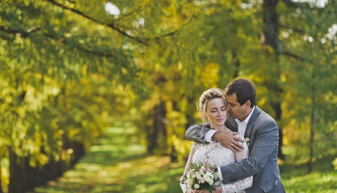 Groom embracing bride from behind in tree-lined garden path during golden hour.