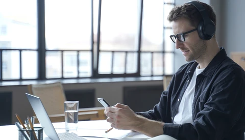 Man wearing headphones at desk working on laptop and smartphone in bright office with city view.