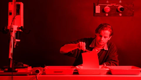 Photographer working in a red-lit darkroom, handling photographic paper under safelight conditions.