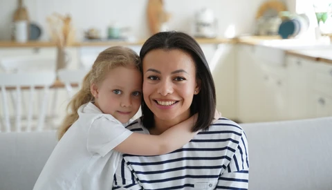 Woman and young child hugging on a couch, both smiling at camera in bright home interior.
