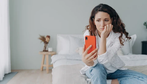 Woman in white shirt holding orange phone while sitting on bed, chin resting on hand.