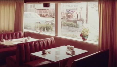 Retro diner booth with burgundy vinyl seating and white Formica table beside large storefront windows.