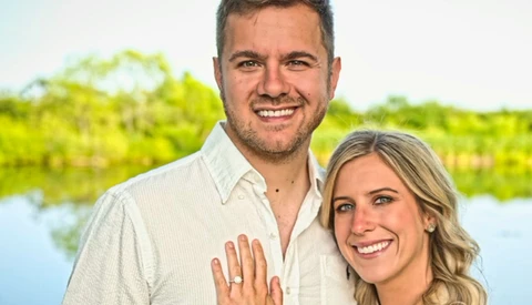 Smiling couple posing outdoors with water and green foliage in soft-focus background.