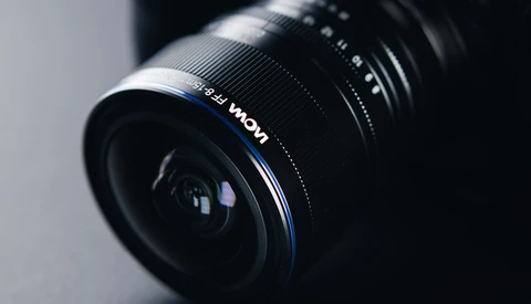 Wide-angle camera lens with blue accent ring mounted at an angle against a neutral gray background.