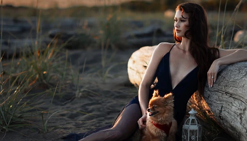 Woman in dark swimsuit seated on driftwood at beach during golden hour, holding small dog.