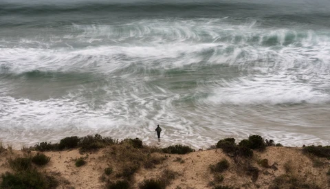 Solitary surfer standing on sandy beach with dune vegetation, facing large rolling waves under overcast sky.