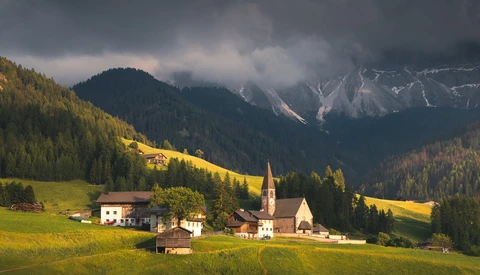 Alpine valley with a small village nestled among forested slopes beneath dramatic snow-capped peaks.