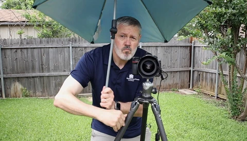 Videographer holding camera on tripod under umbrella in backyard setting.