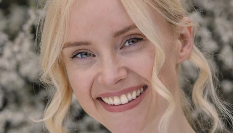 Blonde woman with wavy hair smiling at camera against textured gray background.