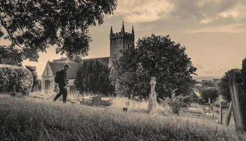 Sepia-toned cemetery scene with stone church and gravestones framed by overhanging trees.