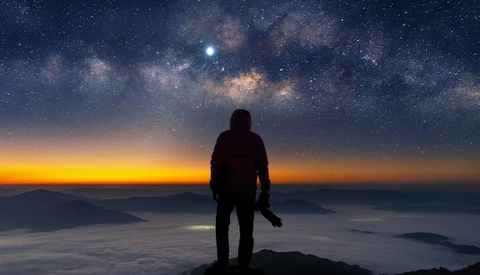 Silhouetted figure standing on a mountain peak beneath the Milky Way galaxy at dawn.