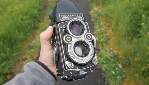 Rolleiflex twin-lens reflex camera held in hand against a blurred green field background.