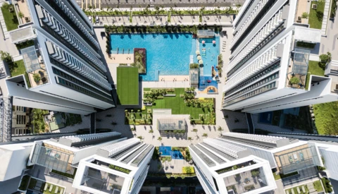Aerial view of a modern residential complex with a bright blue swimming pool surrounded by manicured green spaces and multi-story buildings.