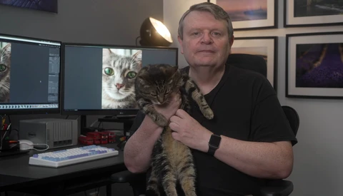Man holding a tabby kitten in a photography studio workspace with dual monitors displaying cat images.