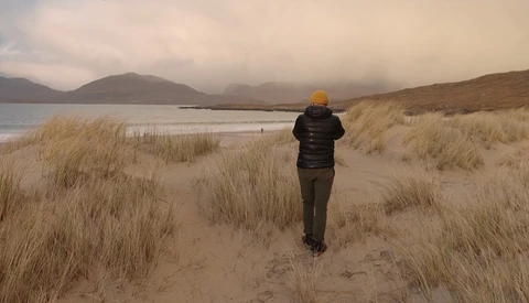 Runner in yellow hat jogging through sandy beach with dune grass toward misty mountains and water.