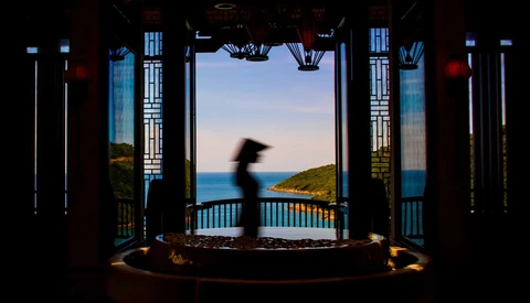 A blurred figure moves through an ornate pavilion overlooking a coastal landscape at twilight.