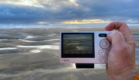 Hand holding a compact digital camera displaying a coastal landscape on its rear screen.