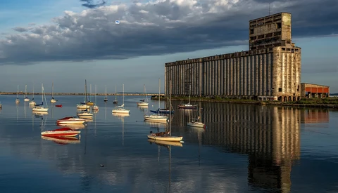 Sailboats moored in calm water with a large industrial grain elevator reflected in the glassy surface at golden hour.