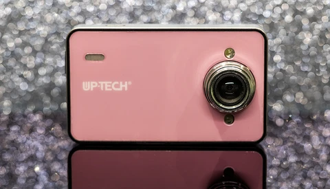 Pink compact digital camera with chrome lens ring against sparkly gray background.