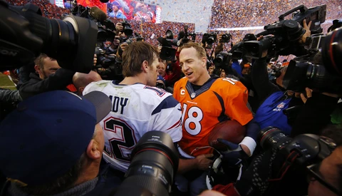 Football player in orange jersey surrounded by photographers and media at stadium field level.