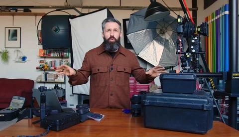 Bearded man in burgundy shirt standing at wooden desk surrounded by photography lighting equipment and cases.