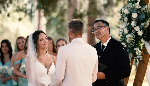 Bride and groom exchange vows at outdoor wedding ceremony with officiant and guests watching.