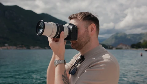 Photographer with telephoto lens photographing across a lake toward mountains on a partly cloudy day.