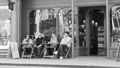 Four people seated outside a storefront with large windows, conversing in black and white.