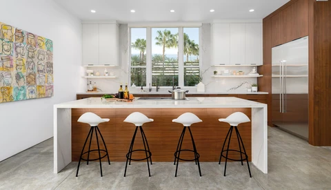Modern kitchen interior with wood and white island featuring four bar stools, stainless steel appliances, and palm tree views through large windows.