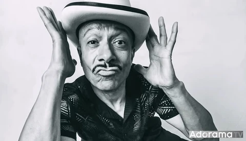 Man in cowboy hat and patterned shirt with hands raised to sides of head, black and white portrait.