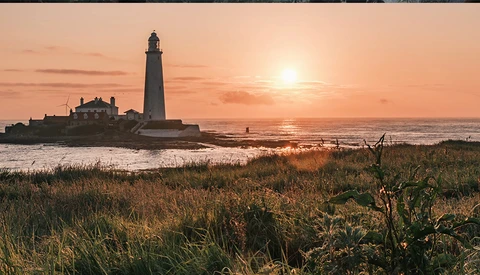 Lighthouse silhouetted against golden sunset sky with keeper's house and coastal grassland in foreground.