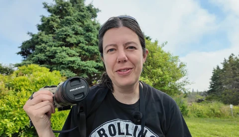 Photographer holding a vintage SLR camera with manual lens, smiling outdoors with trees and green fields in background.