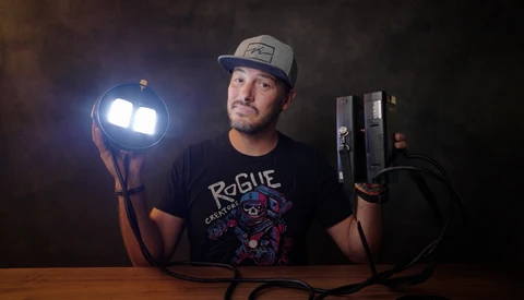 Photographer holding a portable LED light panel in one hand and lighting equipment in the other against a dark background.
