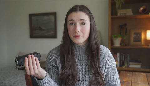 Young woman in gray ribbed sweater holding small handheld device toward camera in home interior.